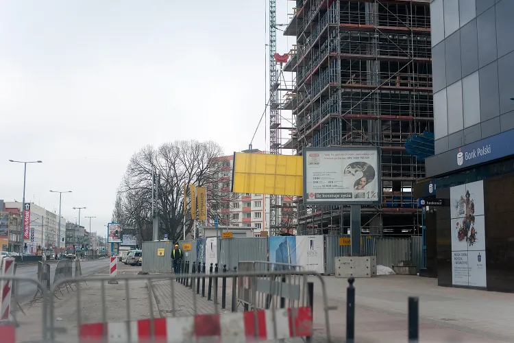 Wjazdu na budowę biurowca we Wrzeszczu pilnuje ochrona, ale - przy tak dużej budowie - ruch ciężarówek jest spory. Tuż za ogrodzeniem dźwig podnosi z ziemi kolejne elementy konstrukcji budynku.  