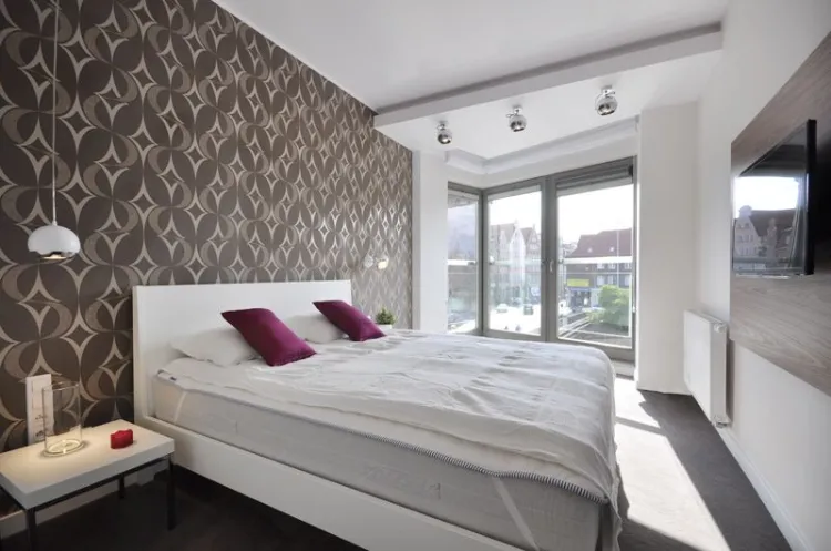 Surowe i nowoczesne wnętrze sypialni apartamentu ocieplone brązową, przytulną tapetą.