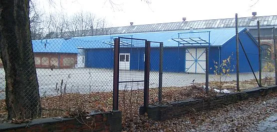Po lewej zabytkowy ceglany budynek pokryty blaszanym, niebieskim dachem. Po prawej nowy barak, postawiony na terenie wpisanym do rejestru zabytków.