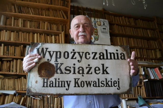 Architekt Janusz Kowalski na tle pozostałości księgozbioru swoich rodziców i z pamiątkowym, metalowym szyldem dawnej wypożyczalni książek.