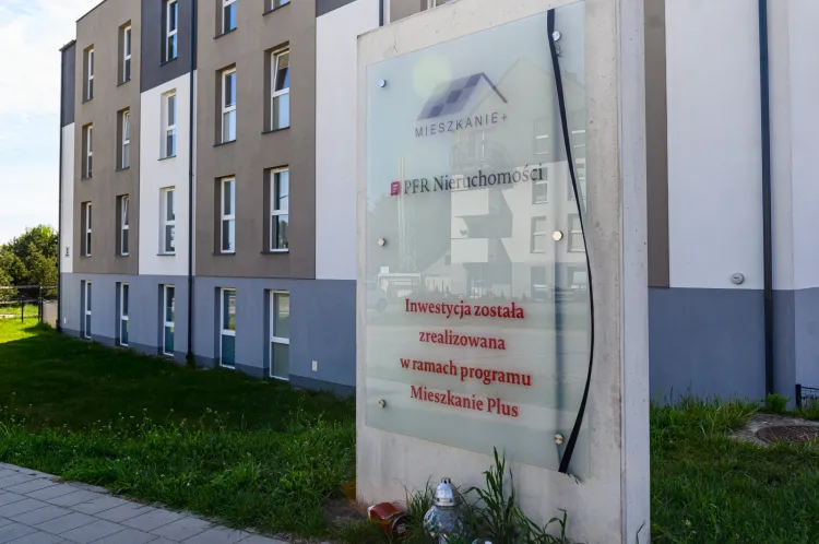 Osiedle Mieszkanie Plus w Gdyni. Tablica informująca, że osiedle powstało w ramach programu przewiązana została kirem. Stanęły pod nią znicze. 